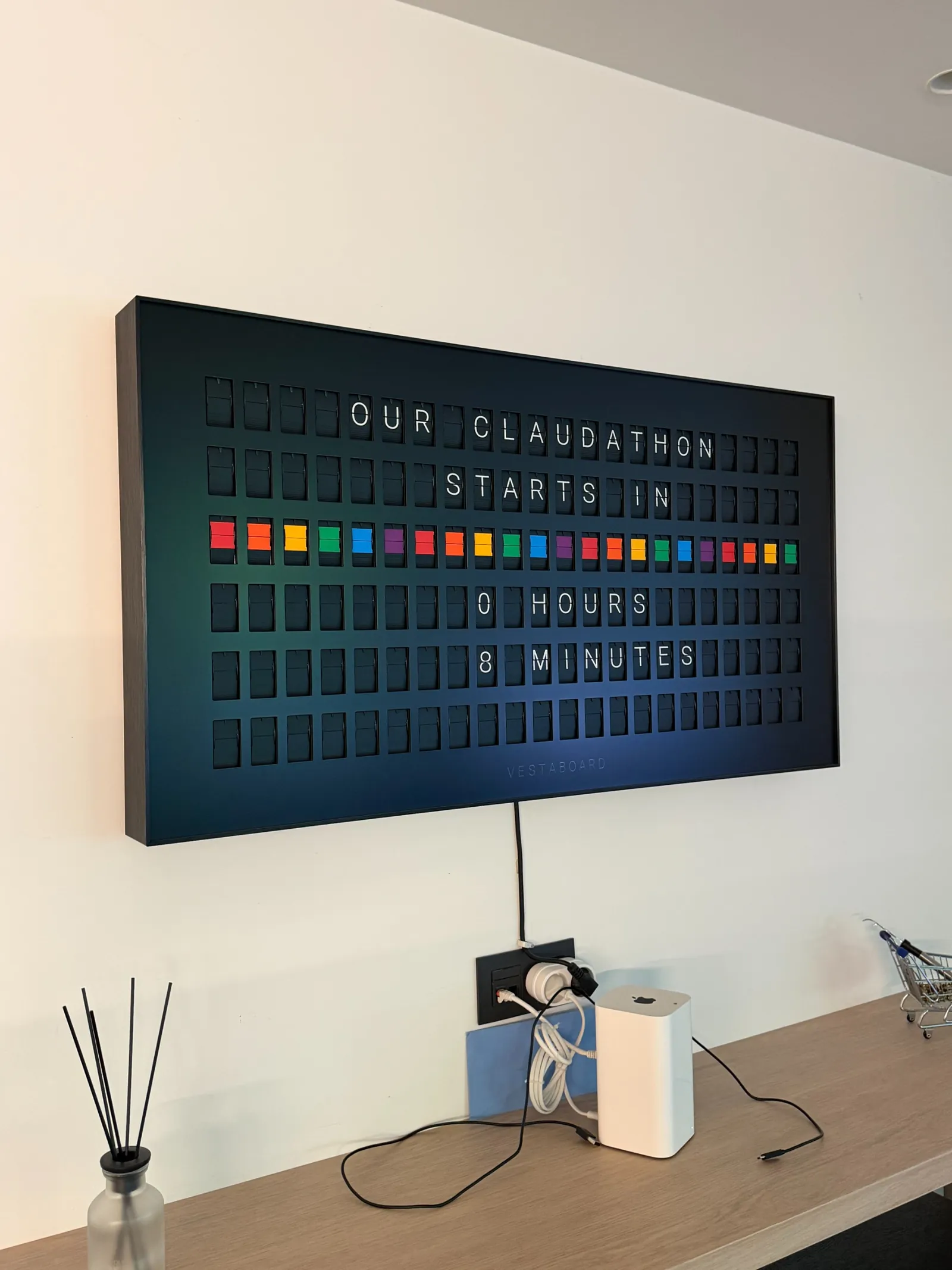 Een digitaal display aan de muur toont met gekleurde blokjes en witte letters "Our Claudathon starts in 0 hours 8 minutes".