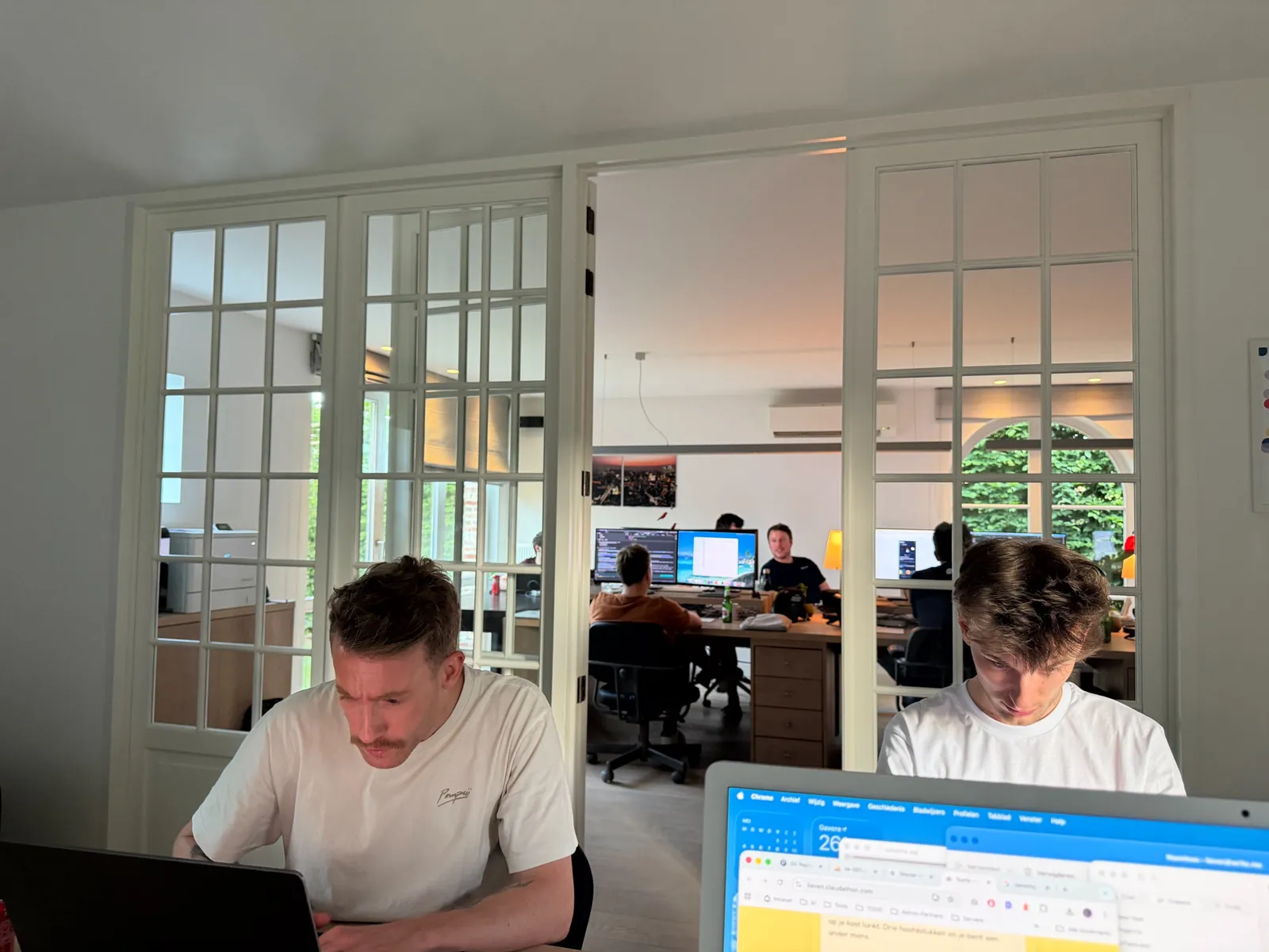 Twee personen werken aan hun bureaus in een modern kantoor, gescheiden door grote glazen deuren met raamwerk, met zicht op andere medewerkers in de achtergrond.